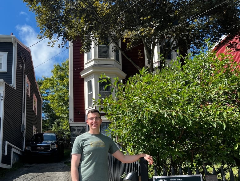 A man stands in the front yard of a house, next to Registered Heritage Structure sign on a black fence.