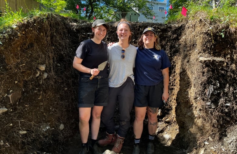 Three people stand arm in arm in a dirt pit the same height as they are, one holding a trowel.