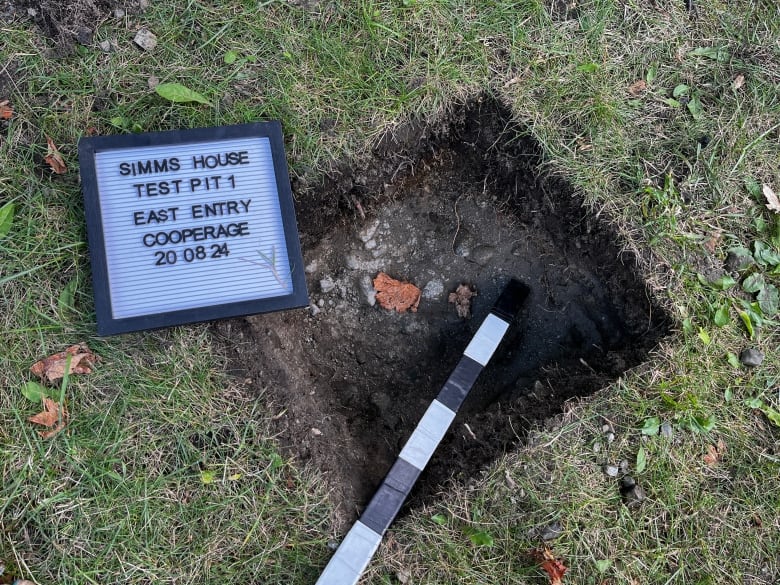 A square hole in the ground surrounded by grass is marked by a sign that reads, 