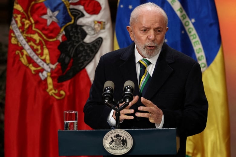 A man speaks at a podium in front of a line of flags, including Brazil's flag. 