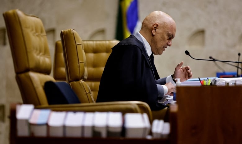 A judge sits at a desk, speaking into a microphone. He is viewed from the side. 