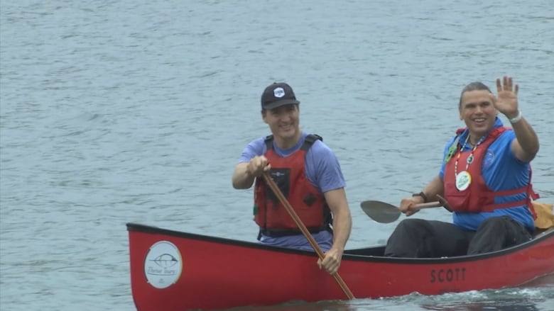 A politician in a blue shirt and an orange life jacket, wearing a black ball cap, paddles in a red canoe with another man in a blue shirt who is waving.