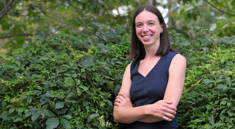 Smiling woman stands with her arms crossed in front of a green foliage background.