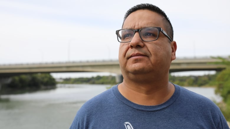 A man with black-rimmed glances looks to the sky, with a riverscape seen behind him.