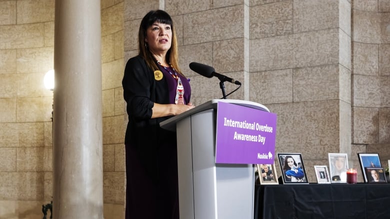 A woman wearing a beaded necklace and a purple shirt speaks at a microphone.