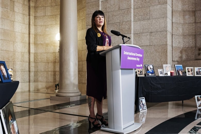 A woman wearing a beaded necklace and a purple shirt speaks at a microphone.