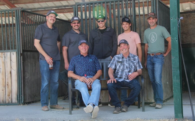 Five men stand in front of two seated men outside empty horse stables.