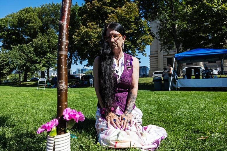 A woman kneels near a tree with flowers at the bottom.