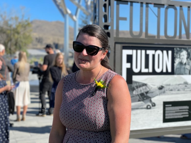 A woman in sunglasses with a yellow flower on the strap of her dress stands with a small crowd of people in Halifax.