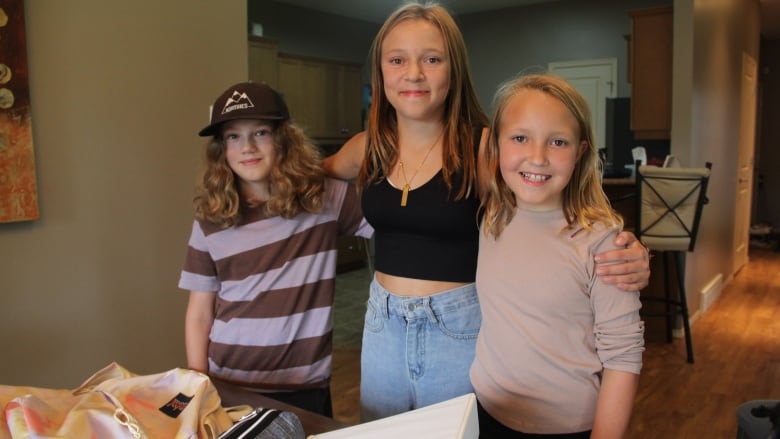 A boy and two girls have their arms around each other, behind a dining room table filled with some of their school supplies.