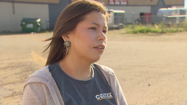 An Indigenous woman with brown hair is wearing a grey zip-up sweater over a charcoal-coloured shirt. She is standing outside in a gravel lot.