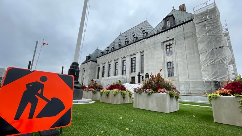 An orange construction sign sits on the green lawn in the foreground of the grey-bricked Supreme Court Building beneath cloudy skies