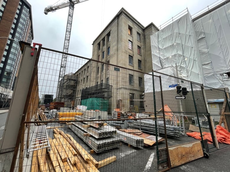 Behind a tall metal fence construction materials are neatly scattered in front of a brown-bricked building. A tall crane keeps watch in the background