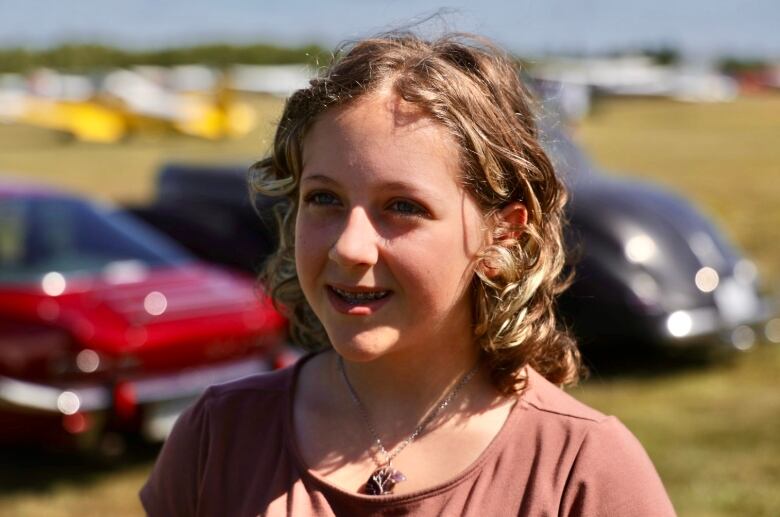 A young girl with braces and curly hair wears a dusty rose coloured shirt. 