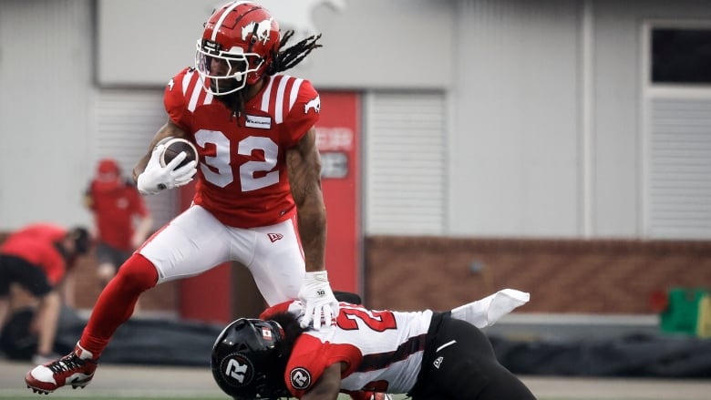 A man in white football uniform with a black helmet tackles a man wearing a red jersey and helmet and white pants on a football field.