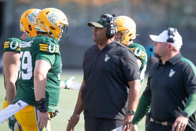 A mean wearing black talks to two men wearing football uniforms and helmets.