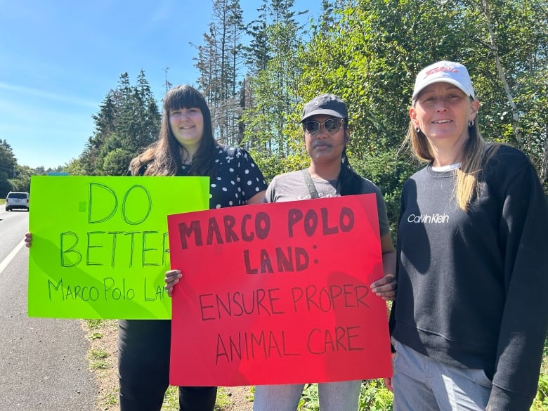 Three women holding protest signs standing on a roadside in a rural area.