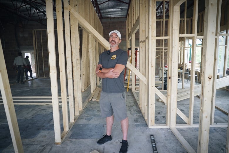 A man in a grey shirt and shorts stands with a smile and his arms crossed as construction is going on inside their newly purchased building. 