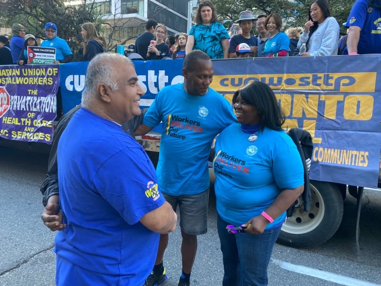 Members of the Canadian Union of Postal Workers, wearing blue union shirts, stand in front of a bus of dozens of union members on a city street in Toronto in daylight,