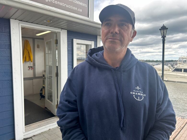 man wearing a cap and navy sweater with the Chambly Marina logo on it standing in front of his offices