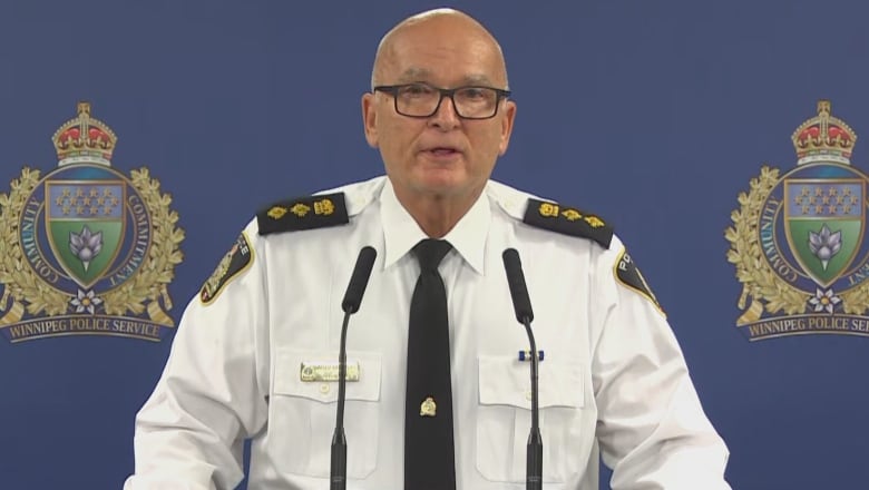 A bald man in a white shirt stands at a podium with microphones. 