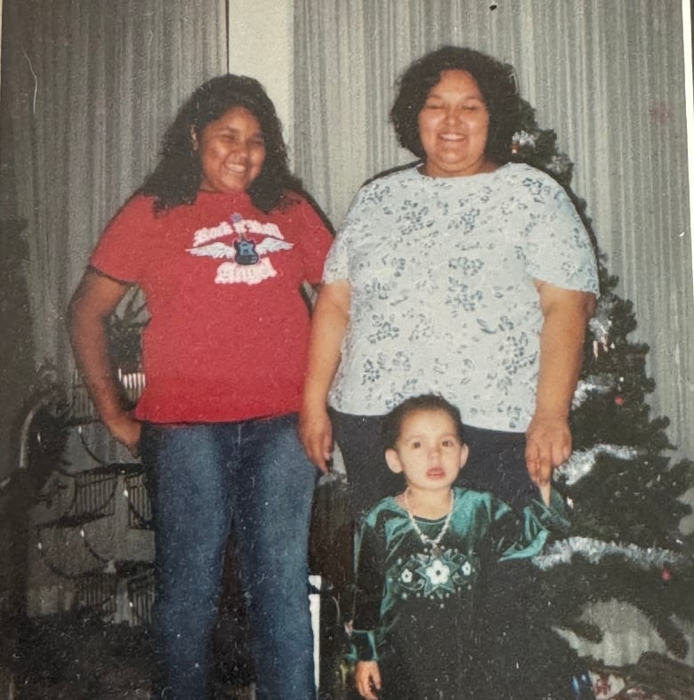 Two adult women stand beside a small Christmas tree, with a girl in a dark green dress standing in front of them. 