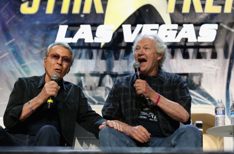 Two men sit beside each other holding microphones while talking.