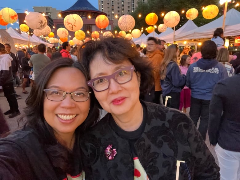 Two women pose for a photo with hundreds of lit lanterns hanging above the street behind them. 