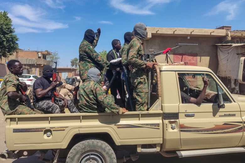 Men in camouflage uniforms in the back for a pick-up truck. One man is standing behind a cab holding an automatic weapon. 