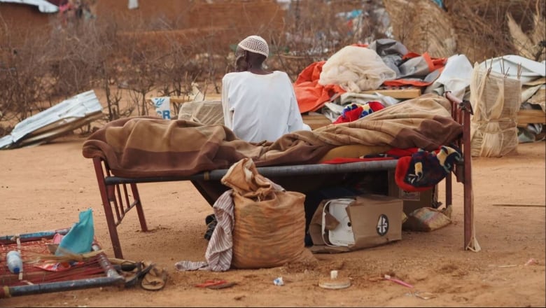 A man wearing a white garment and white hat on his head sits with his back turned, next to his belongings, on a bed that is outside on dirt. 