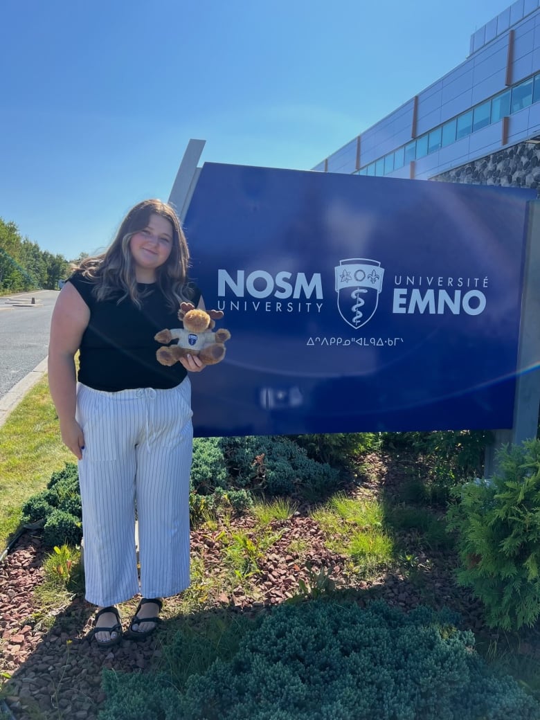 A young woman holding a teddy bear and standing in front of a sign that says NOSM.