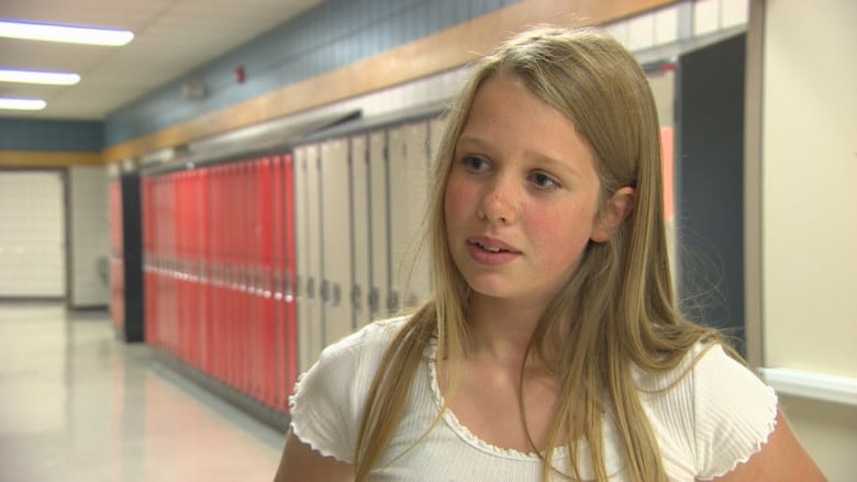 A young girl in long blonde hair stands in a school hallway.