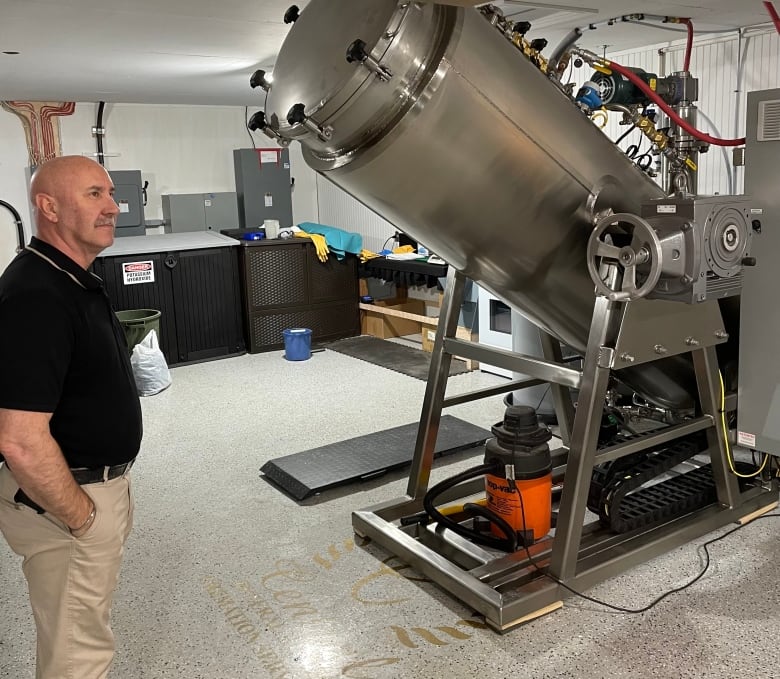 Man in black polo shirt and khaki pants standing next to a machine that has a cylindrical tube held at an angle.