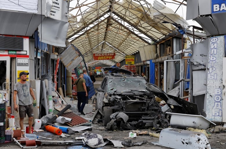 Debris is shown around and on top of a heavily damaged vehicle on a narrow laneway as a few individuals are shown standing about.