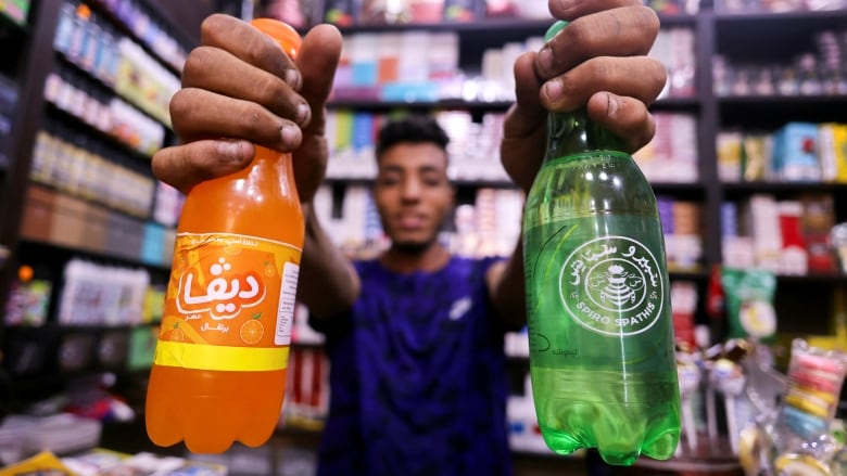 A man holds two bottles of different brands of soda up to the camera. 