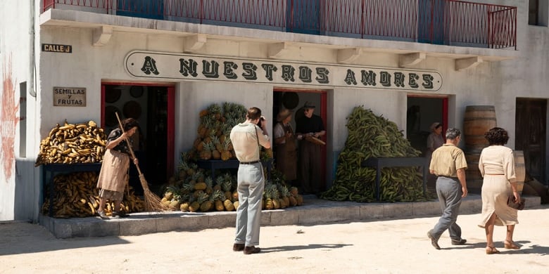 outside a produce store in Mexico City, a woman sweeps the curb, people talk in the background as a man takes a photo.