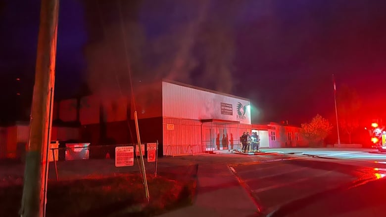 A red-tinged pall envelops a squat, square building surrounded by first responders at night.