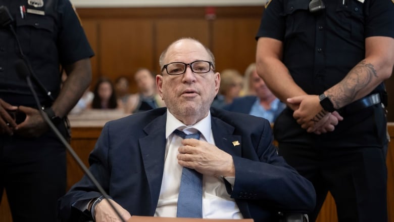 An older, balding bespectacled man in a suit and tie sits at a table in a courtroom.