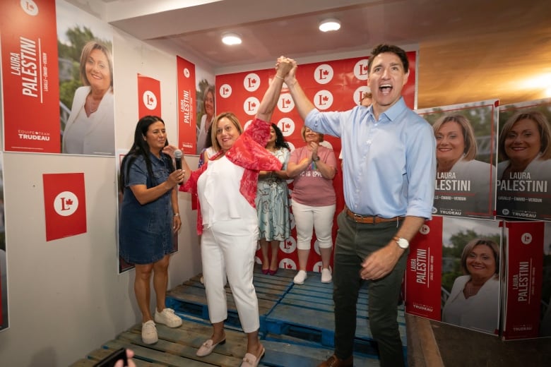 A man in a blue button up shirt raises the hand of a woman wearing a white shirt and red overshirt. People in the background applaud.