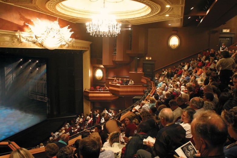 People sit in an old-fashioned theatre with loges and chandeliers, holding programs. 