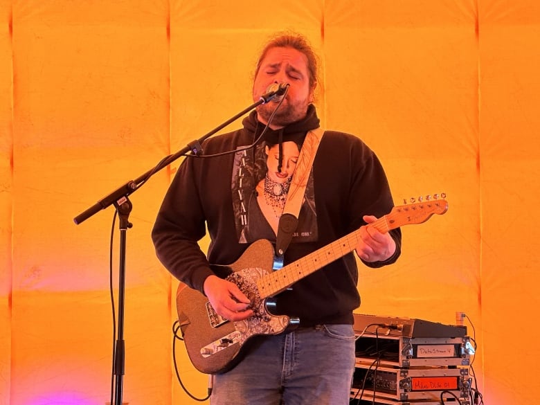 A person sings into a microphone and strums a guitar in an orange-lit room.