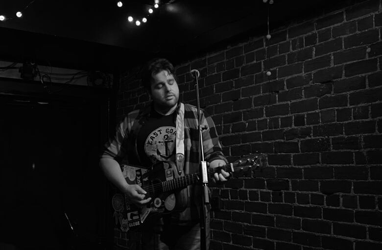 A person wearing a flannel shirt sings into a microphone while strumming a guitar in front of a brick wall.