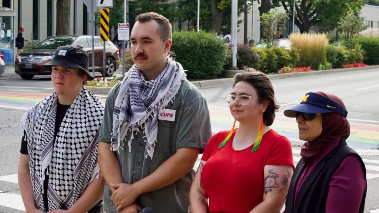 Four people stand in a line at a city street corner with their backs to the road. 