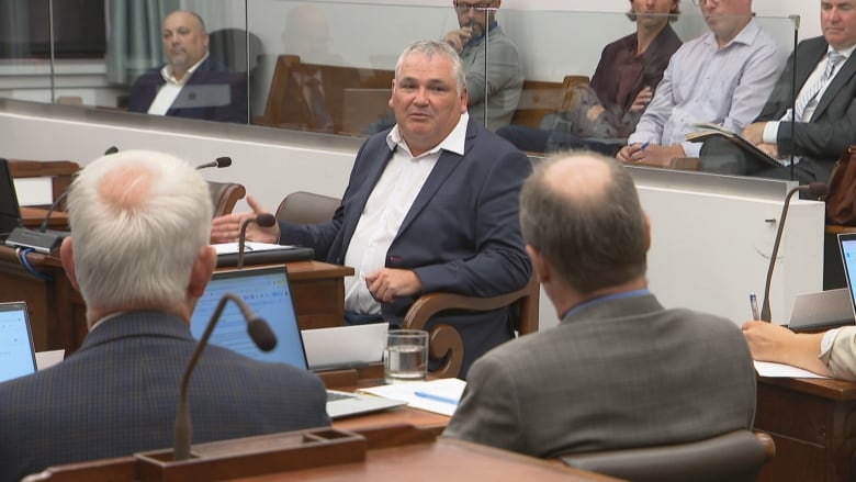 A man in a dark blue suit and grey hair sits in a legislative assembly. 