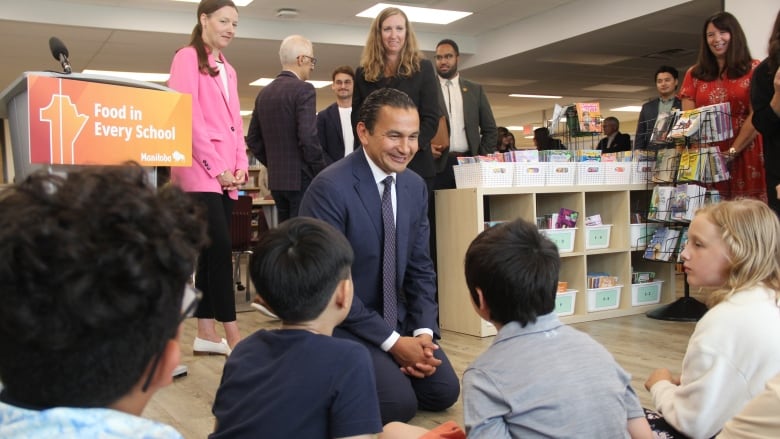 A man in a dark blue suit and tie kneels on the floor in front of a number of kids sitting on a rug.