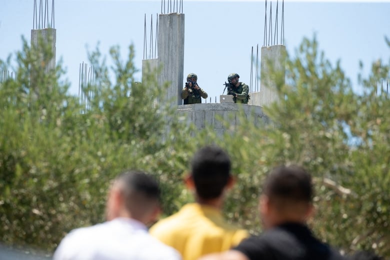 A few people are shown in the foreground, blurred, as farther away two military members in gear with weapons pointed are shown from a rooftop perch.