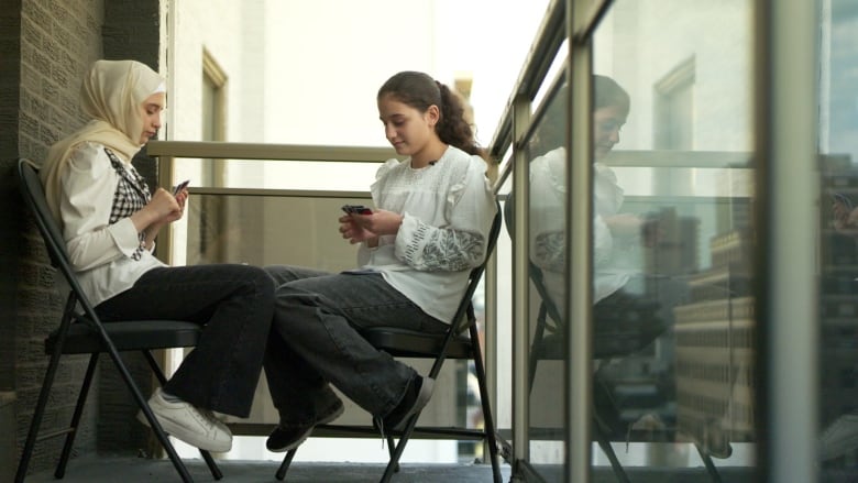 Two girls sitting on chairs in the corner of a balcony. The girls are sitting face to face, their knees touching, while they play the UNO card game.