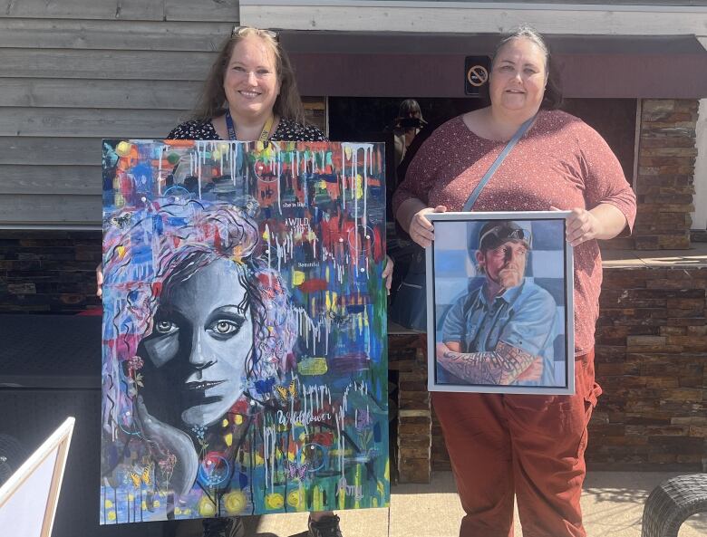 two women stand side-by-side holding portraits of people