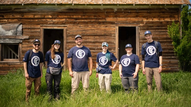 Six people standing in front of an abandoned building. They are all wearing matching navy blue shirts with a logo of the Mtis Nation of Alberta on it. 