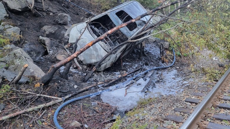 Scene from a vehicle crash, showing a burned car after collision with a tree, damaged tree trunks, and shattered glass on the ground by a rail track. 